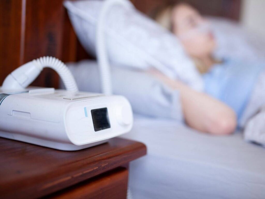 A CPAP machine beside a sleeping woman highlights the life-saving role standby generators play during power outages for medically vulnerable families.